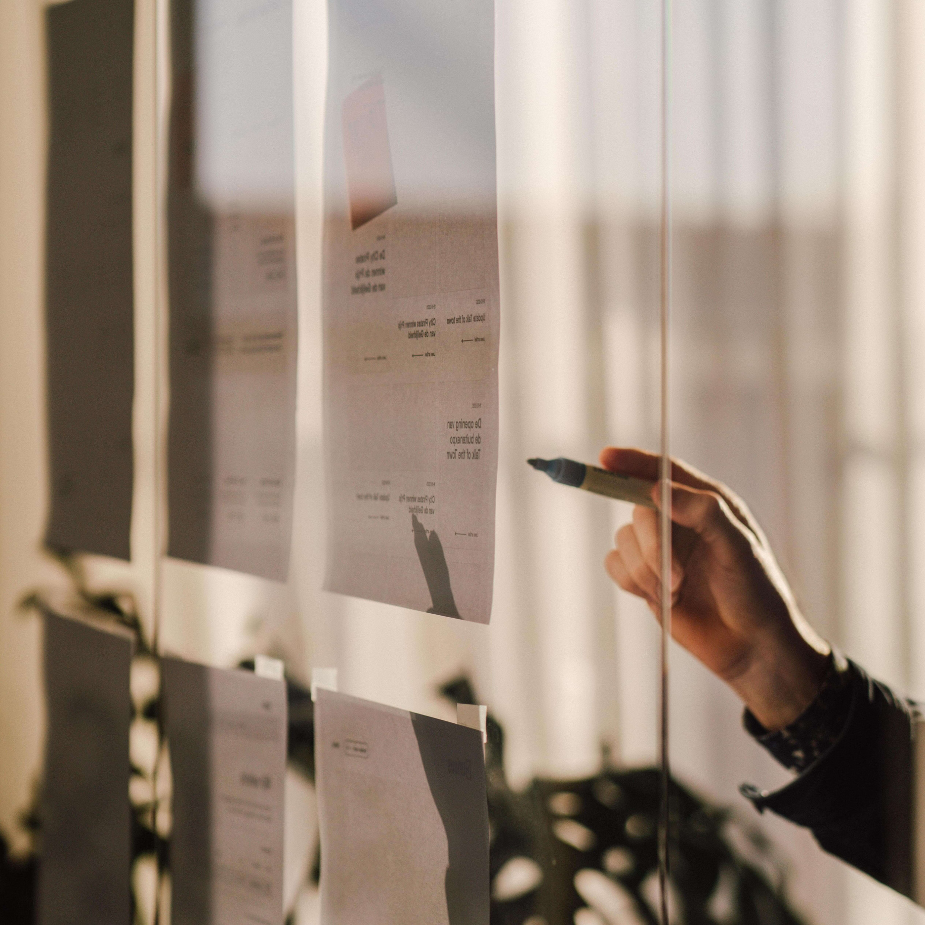 Hand pointing with a marker at printed design layouts taped to a glass partition in a sunlit office