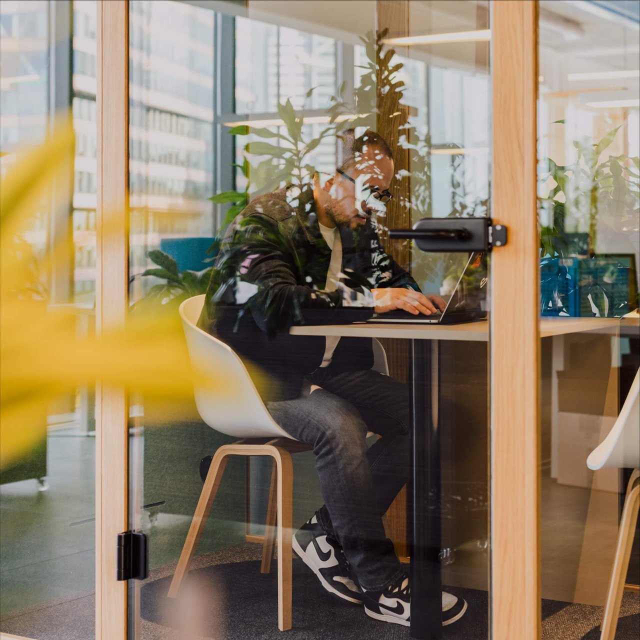 Man working on a laptop in a glass-walled Bynder's office pod, seen through glass with reflections of plants and city buildings, in a bright, plant-filled modern workspace.