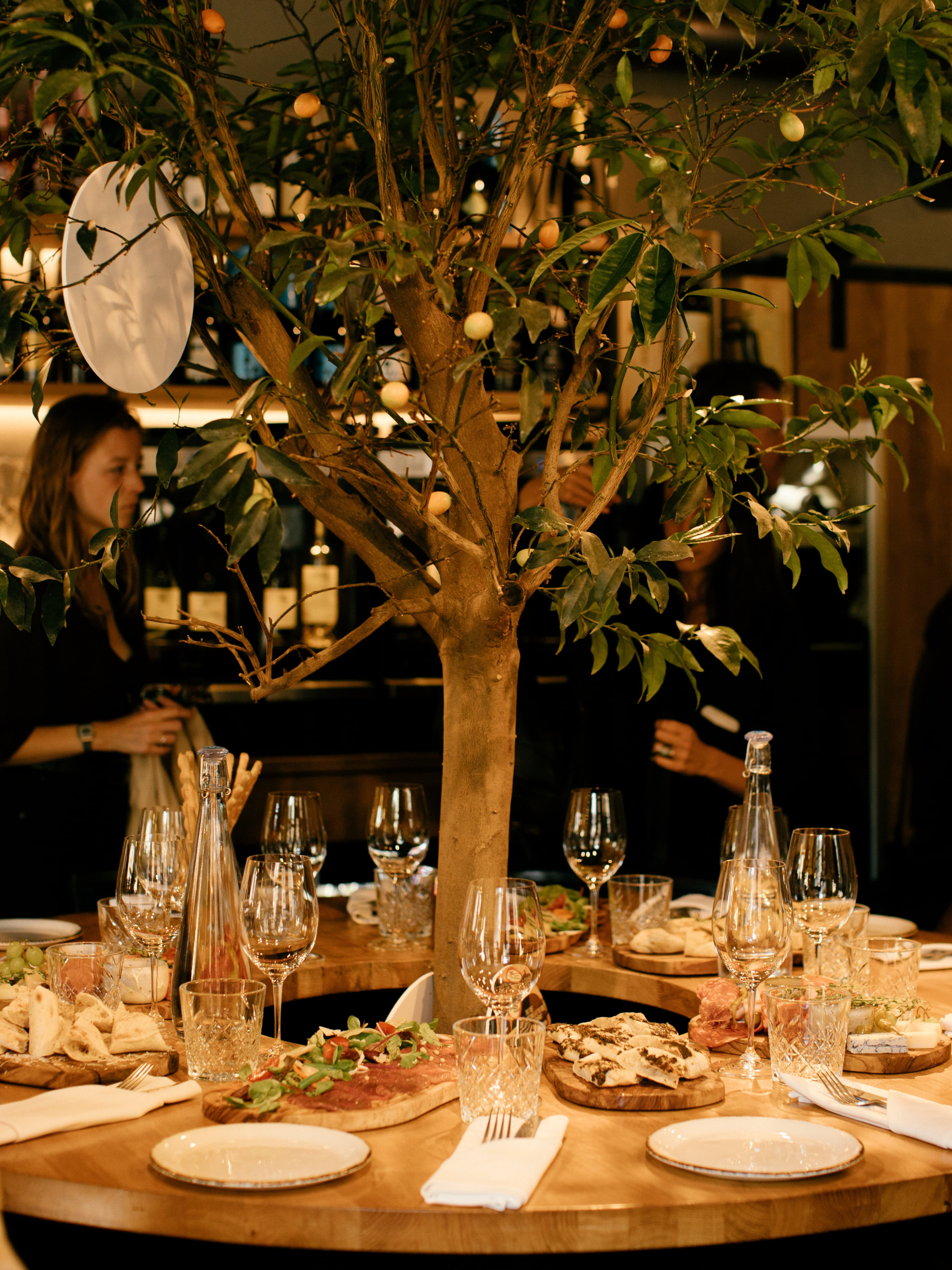 Circular wooden table featuring a central citrus tree, wine glasses, and shared food platters in Tallinn