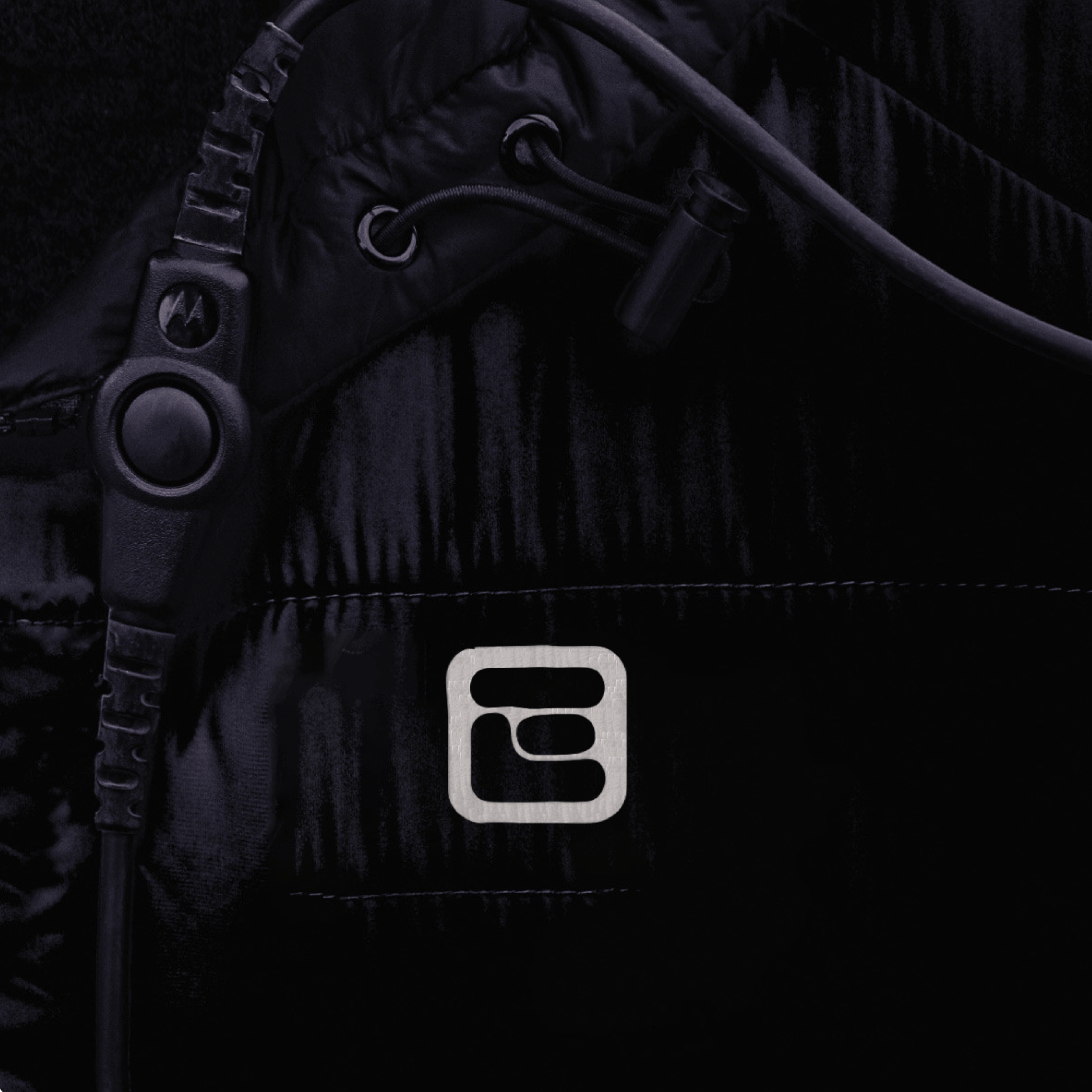 Close-up of a black puffer jacket with the ECommpay silver logo icon on the chest and a radio earpiece cable clipped to the front.